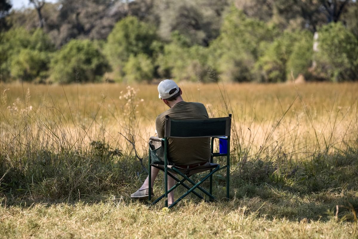 Een deelnemer zit alleen in een kampeerstoel en kijkt uit over het goudkleurige gras van de Okavango Delta
